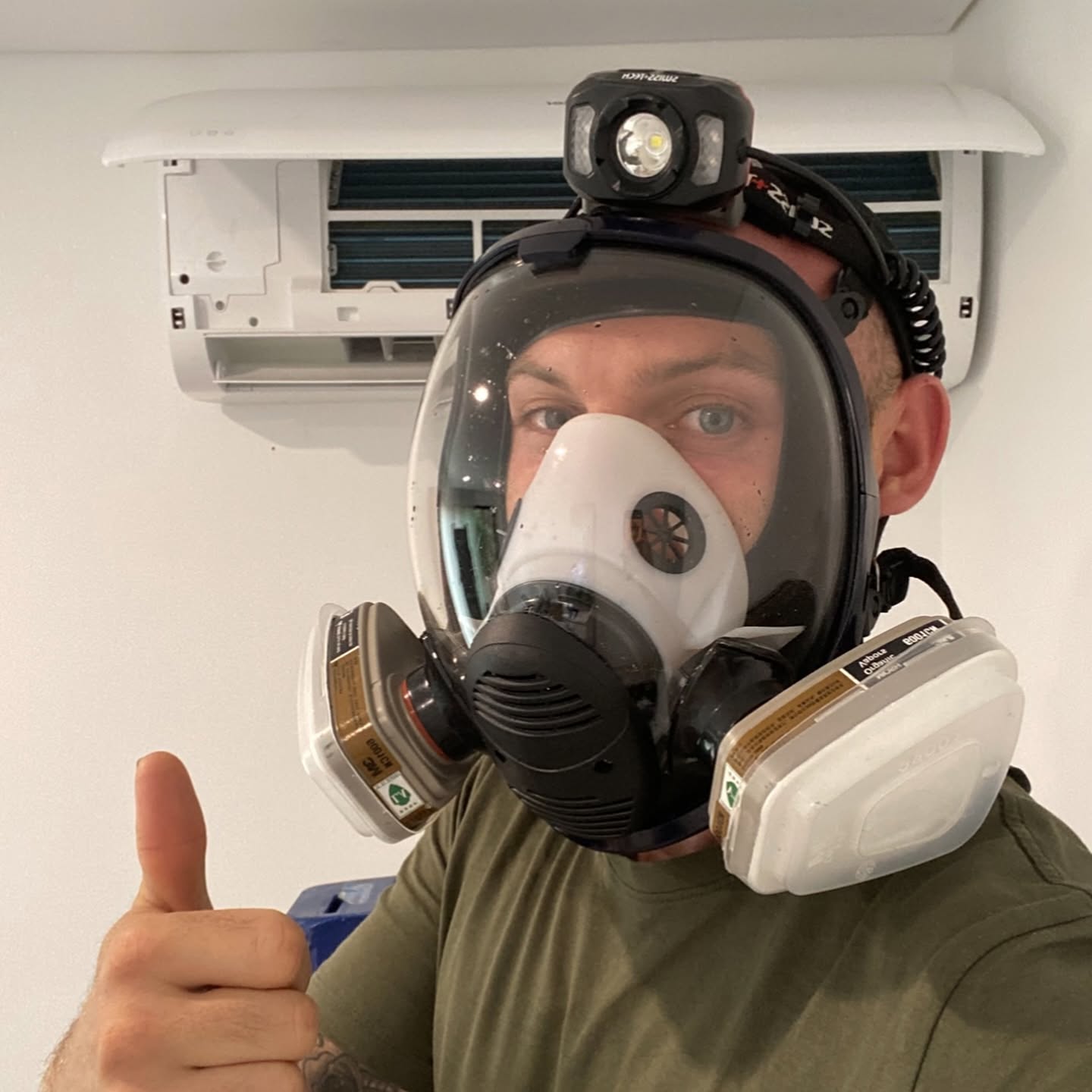 Natural Breeze technician in a full-face respirator and headlamp, thumbs up in front of a split system